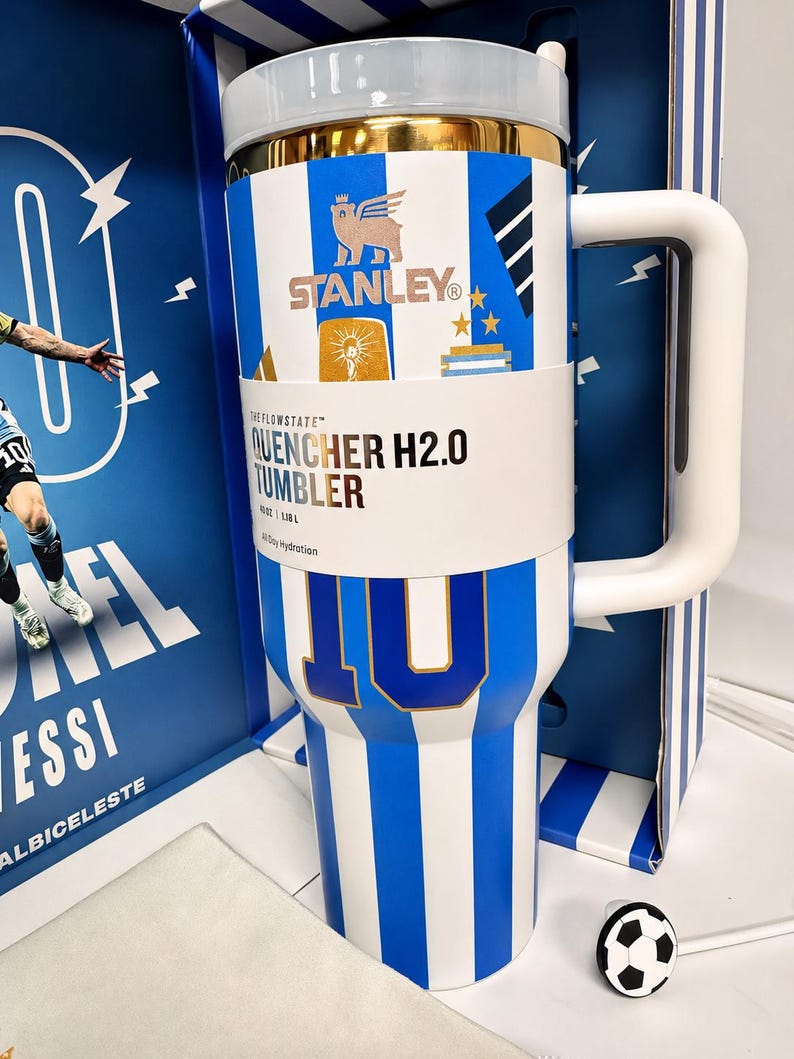 May include: A white and blue striped Stanley Quencher H2.0 tumbler featuring a gold rim, a large white handle, and a paper band with the text The Flowstate Quencher H2.0 Tumbler 40 oz 1.18 L. The tumbler displays the number 10 and a soccer ball charm.