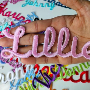May include: A pink satin embroidered patch with the name "Lillie" in a cursive font.