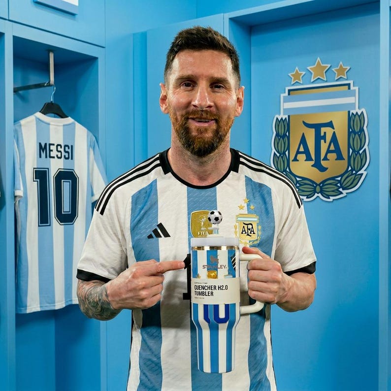 May include: A white and light blue striped Stanley Quencher H2.0 tumbler featuring a soccer ball topper on the lid. The tumbler has a large handle and a paper label with the text Stanley and Quencher H2.0 Tumbler printed on the front surface.