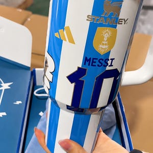 May include: A white and blue striped insulated tumbler featuring the Stanley logo, a gold emblem, the name MESSI, and the number 10 in dark blue. The design includes a gold three-stripe logo and a white handle, with a clear lid at the top.