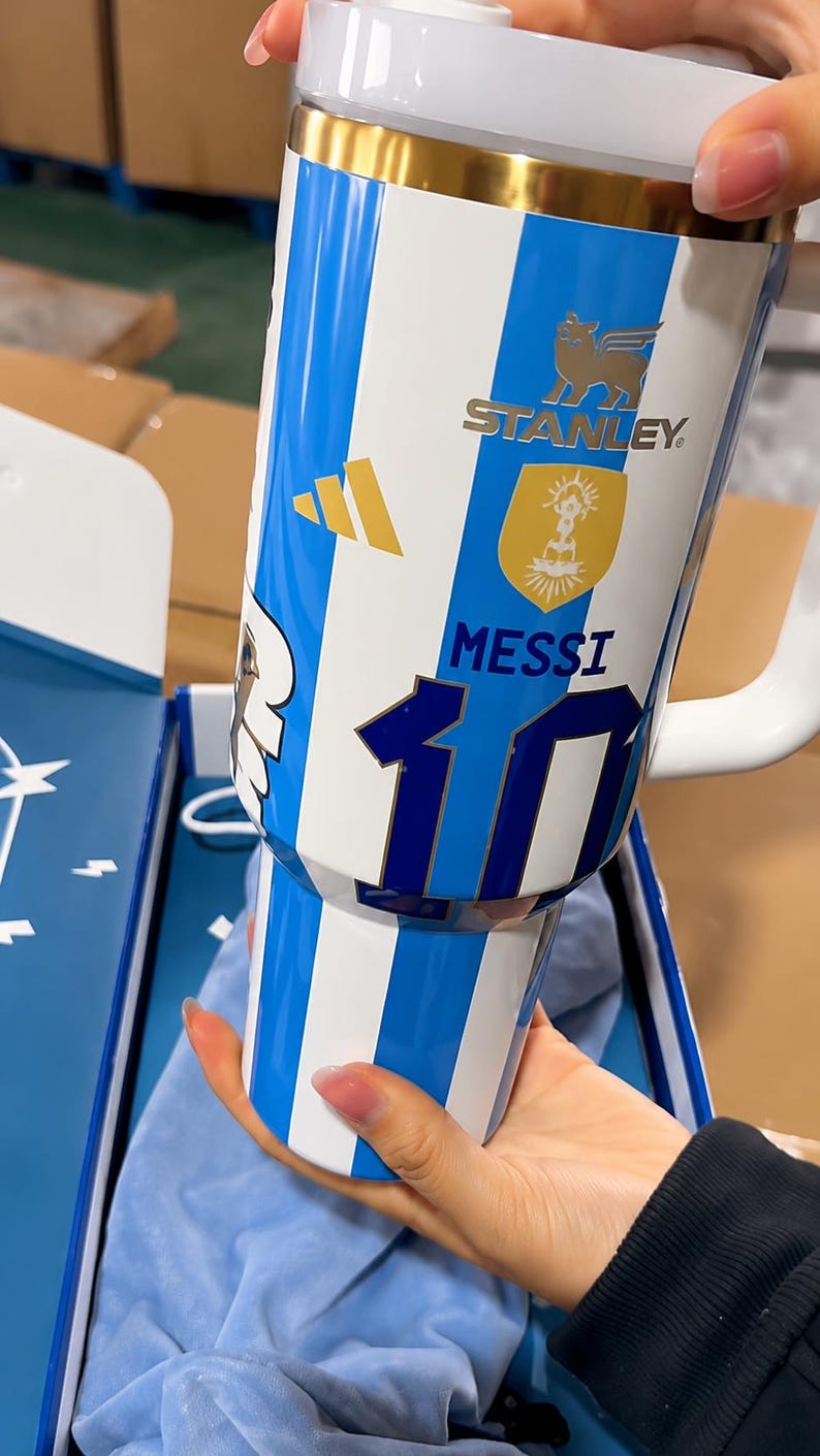 May include: A white and blue striped insulated tumbler featuring the Stanley logo, a gold emblem, the name MESSI, and the number 10 in dark blue. The design includes a gold three-stripe logo and a white handle, with a clear lid at the top.