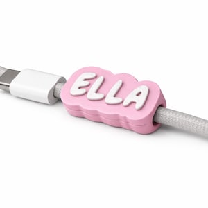 Peut inclure: Un organiseur de câble rose avec le nom "ELLA" en lettres blanches. L'organisateur est fixé à un connecteur USB-C blanc et à un câble tressé gris. Le design est simple et moderne, adapté à l'organisation des câbles de chargement.