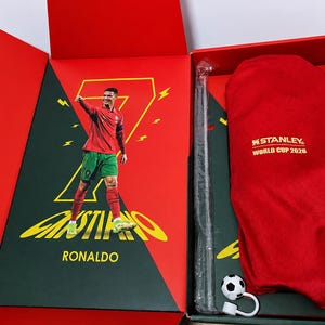 May include: A red and green gift box featuring a graphic of Cristiano Ronaldo in a soccer uniform with the number 7. Inside is a red fabric cover with the text Stanley World Cup 2026, a long thin black object in plastic, and a small soccer ball charm.