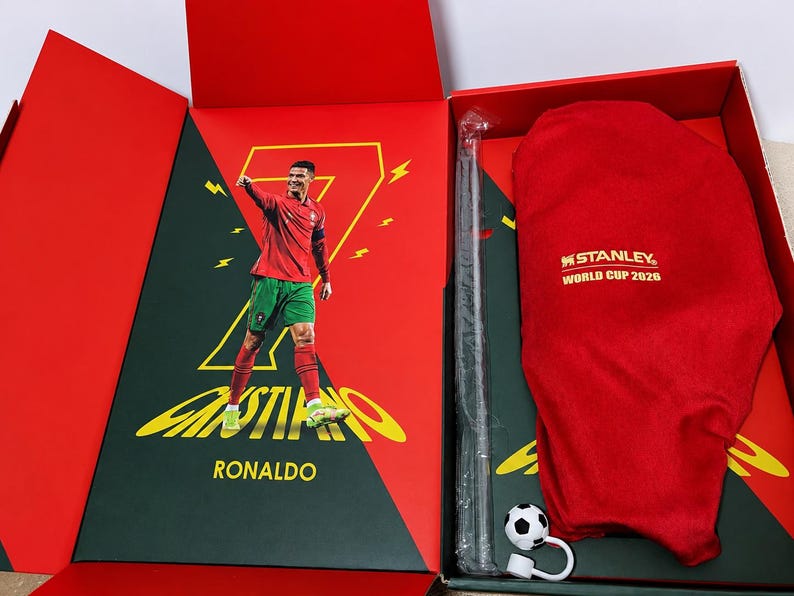 May include: A red and green gift box featuring a graphic of Cristiano Ronaldo in a soccer uniform with the number 7. Inside is a red fabric cover with the text Stanley World Cup 2026, a long thin black object in plastic, and a small soccer ball charm.