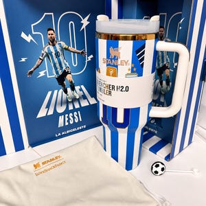 May include: A white and blue striped Stanley Quencher H2.0 tumbler featuring the number 10, a gold rim, and a white handle. It is presented in a matching box with a Lionel Messi graphic and includes a cream-colored drawstring bag with gold text.