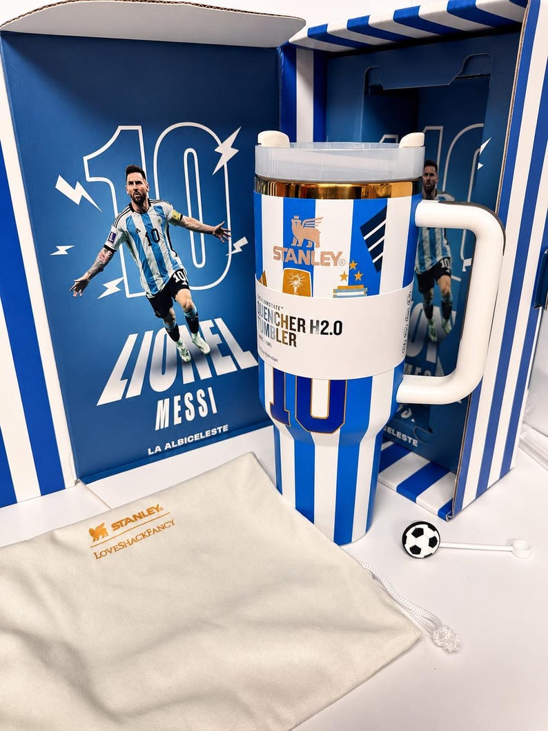 May include: A white and blue striped Stanley Quencher H2.0 tumbler featuring the number 10, a gold rim, and a white handle. It is presented in a matching box with a Lionel Messi graphic and includes a cream-colored drawstring bag with gold text.