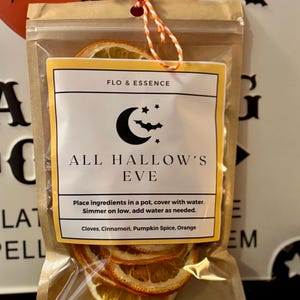 May include: A clear plastic bag with dried orange slices and a label that reads "FLO & ESSENCE" and "ALL HALLOW'S EVE". The label also includes instructions and lists ingredients: cloves, cinnamon, pumpkin spice, and orange. The bag is tied with orange and white twine.