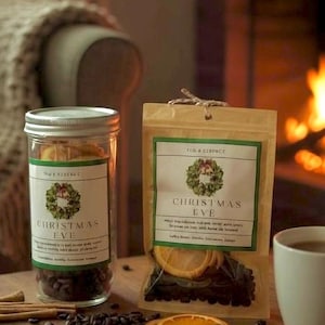 May include: A clear glass jar and a tan pouch labeled "Christmas Eve" with a wreath design. The jar contains dark beans, and the pouch holds dried orange slices. Cinnamon sticks and coffee beans are scattered on a wooden surface.