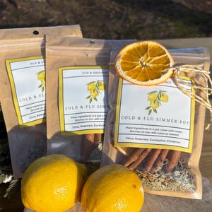 Op de afbeelding: Drie bruine papieren zakjes met een wit label waarop "Cold & Flu Simmer Pot" staat. Het label heeft een afbeelding van citroenen en de tekst "Place ingredients in a pot, cover with water. Simmer on low, add water as needed. Citrus, Rosemary, Eucalyptus, Cinnamon". De zakjes bevatten kaneelstokjes, gedroogde sinaasappelschijfjes en andere ingredi&euml;nten.