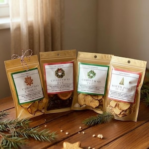 May include: Four clear cellophane bags filled with dried fruit and spices, labeled "Christmas Cookies", "Christmas Eve", "Christmas Tee", and "North Pole". The bags are on a wooden surface with a star-shaped cookie and pine sprigs.