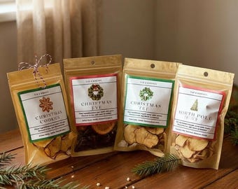 May include: Four stand-up pouches with clear windows containing dried fruit slices. Labels read Christmas Cookies, Christmas Eve, Christmas Tea, and North Pole Tea. Each features a festive graphic and small text. A star-shaped cookie sits in front.