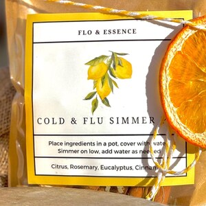 Op de afbeelding: Een geel en wit label met de tekst "FLO & ESSENCE COLD & FLU SIMMER" en instructies voor het maken van een drankje tegen verkoudheid en griep. Het label is versierd met een handgetekende illustratie van citroenen en een gedroogde sinaasappelschijf.
