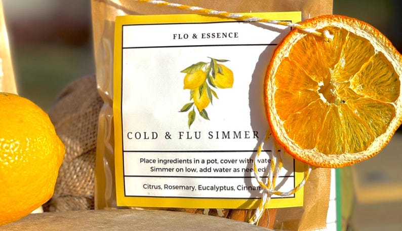 Op de afbeelding: Een geel en wit label met de tekst "FLO & ESSENCE COLD & FLU SIMMER" en instructies voor het maken van een drankje tegen verkoudheid en griep. Het label is versierd met een handgetekende illustratie van citroenen en een gedroogde sinaasappelschijf.