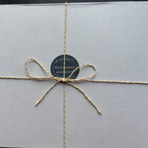Op de afbeelding: Een witte doos met een strik van gele en witte touw. De doos heeft een zwart-wit rond etiket met de tekst "Flo & essence" en het websiteadres "www.floandessence.com".