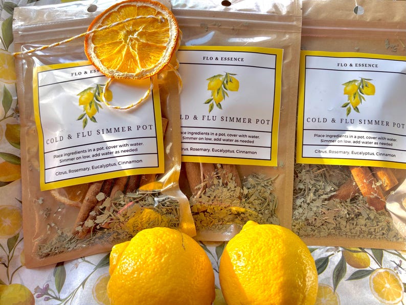 Op de afbeelding: Drie doorzichtige plastic zakjes gevuld met gedroogde kruiden en specerijen voor een stoofpotje tegen verkoudheid en griep. De zakjes zijn gelabeld met "Cold & Flu Simmer Pot" en vermelden de ingredi&euml;nten: Citrus, Rozemarijn, Eucalyptus, Kaneel. Er zijn twee citroenen en een gedroogde sinaasappelschijf op de afbeelding.