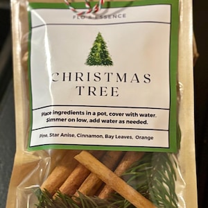 May include: A clear plastic bag containing Christmas tree ingredients. The bag has a label with a green border and the words "CHRISTMAS TREE" and "FLO & ESSENCE". Inside are cinnamon sticks, pine needles, and other ingredients.
