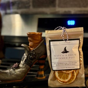 May include: A decorative witch's boot and a bag of Witch's Brew Simmer Pot ingredients. The boot is gray, orange, and black with a spiderweb design. The bag contains dried orange slices and has text that reads "Witch's Brew Simmer Pot."
