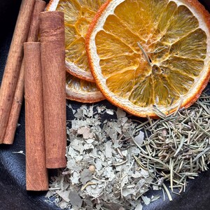 Op de afbeelding: Gedroogde sinaasappelschijfjes, kaneelstokjes, eucalyptusbladeren en rozemarijntakjes in een zwarte gietijzeren pan. Deze ingredi&euml;nten worden vaak gebruikt voor potpourri of om te sudderen op het fornuis voor een natuurlijke geur in huis.