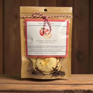May include: A clear plastic bag containing a warm apple pie simmer pot kit. The kit includes dried apple slices, cloves, and cinnamon sticks. A label with the text "WARM APPLE PIE SIMMER POT" is attached to the bag. The bag is tied with a red and white string.