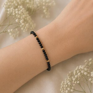 May include: A delicate beaded bracelet featuring small, faceted black stones arranged in a continuous line, interspersed with tiny, smooth, polished gold-toned metal beads that provide a subtle contrast to the dark, reflective surface of the stones.