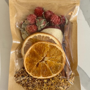 Op de afbeelding: Een doorzichtige plastic zak gevuld met gedroogd fruit, specerijen en kruiden om een winterdrankje te maken. De zak bevat gedroogde sinaasappelschijfjes, kaneelstokjes, gedroogde appelstukjes en rode bloemknoppen.