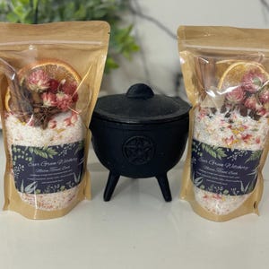 Pode incluir: Duas embalagens de papel kraft cheias de sais de banho, flores secas e rodelas de laranja, com um caldeirão de ferro fundido preto no centro. As embalagens têm rótulos que dizem "Ever Green Witchery".