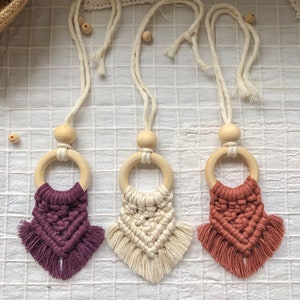 Könnte beinhalten: Drei Makramee-Wandbehänge mit Holzringen und Quasten. Die Behänge sind in den Farben Lila, Creme und Orange.