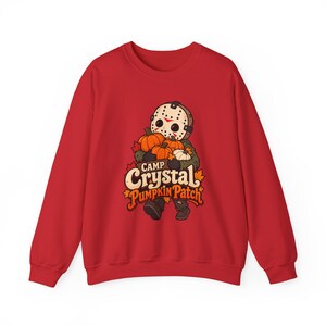May include: A red long-sleeve sweatshirt featuring a graphic of a character wearing a hockey mask and holding orange pumpkins. The text reads Camp Crystal Pumpkin Patch, accompanied by orange and yellow autumn maple leaf illustrations.