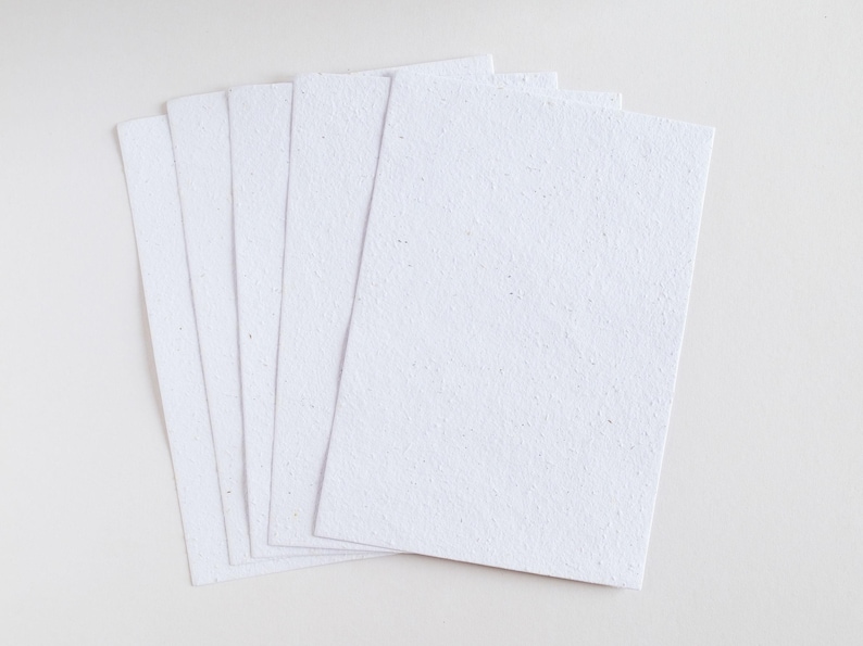 May include: A stack of six rectangular, white paper sheets with a textured surface. The paper is arranged in a slightly fanned-out pattern, showcasing the individual sheets. The background is a plain white surface.
