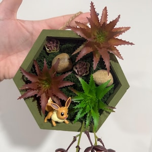 May include: A hexagonal green wooden frame with a moss background and artificial succulents. A small brown and yellow Pokemon figurine is in the center of the frame. A trailing vine with dark purple leaves hangs from the bottom of the frame.
