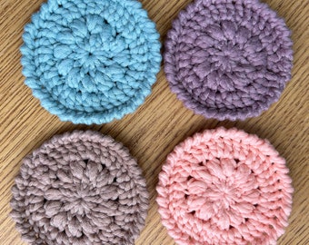 Op de afbeelding: Vier handgemaakte gehaakte onderzetters in tinten blauw, paars, taupe en roze. Elke onderzetter heeft een cirkelvorm met een bloemmotief in het midden. De onderzetters zijn gemaakt van zacht garen.