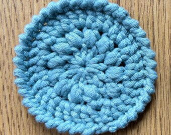 Op de afbeelding: Een ronde, lichtblauwe gehaakte onderzetter. De onderzetter is gemaakt van zacht garen en heeft een getextureerd, verhoogd patroon. De randen zijn netjes afgewerkt. Het meet ongeveer 10 cm in diameter en beschermt oppervlakken.