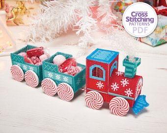 May include: A red and blue toy train with candy cane wheels and two teal boxcars filled with colorful candies. The train is decorated with snowflakes and the text "The World of Cross Stitching Patterns PDF".
