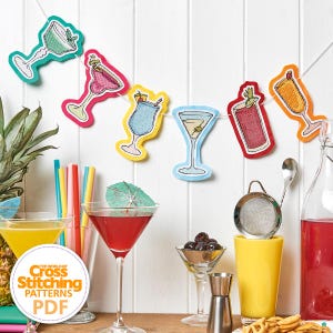 May include: A festive garland of cocktail-themed cross-stitch patterns hangs against a white background. The garland features colorful cocktail glasses in various designs. The image also includes cocktail glasses, a pineapple, and the text "The World of Cross Stitching Patterns PDF".