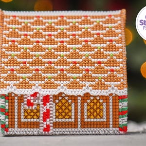 May include: A cross stitch pattern of a gingerbread house with a white roof, red candy canes, and yellow windows. The pattern is on a white background with the text "The World of Cross Stitching Patterns PDF" in a purple circle.