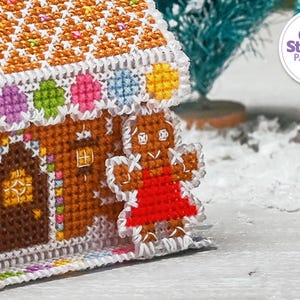 May include: A gingerbread house cross stitch pattern with a gingerbread person standing in front of it. The house is decorated with colorful candies and the gingerbread person is wearing a red dress. The pattern is from The World of Cross Stitching Patterns PDF.