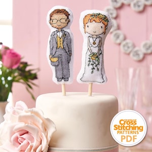 Pode incluir: Um bolo de casamento branco coberto com duas figuras de ponto cruz: um noivo de fato cinzento e uma noiva de vestido branco. As figuras estão em palitos de madeira. O fundo é rosa.