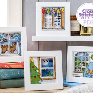 May include: Four white picture frames with cross-stitch designs of winter scenes. The frames are on a shelf with a stack of books and a white blanket.