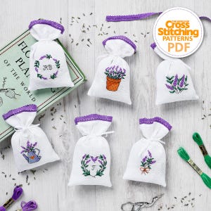 Könnte beinhalten: Weiße Säckchen mit gestickten Lavendelmotiven und lila Spitzenbesatz. Die Säckchen zeigen verschiedene Lavendel-Motive, darunter eine Topfpflanze, eine Gießkanne und Blumenarrangements. Das Bild enthält auch Stickgarn, eine Schere und ein Buch mit dem Titel "FLORA PLANT OF THE WORLD".