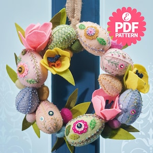 Puede incluir: Una corona de Pascua hecha a mano con huevos y flores de fieltro en colores pastel. La corona incluye flores de fieltro rosas, amarillas y azules, y huevos decorados con bordados y abalorios. La corona está adornada con hojas de fieltro verdes y un colgador de arpillera.