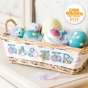 Puede incluir: Una cesta de mimbre llena de huevos de Pascua decorados y plumas. La cesta tiene una banda de tela cosida con la palabra "EASTER" en punto de cruz colorido. Un logotipo circular en la esquina superior derecha dice "The World of Cross Stitching Patterns PDF".