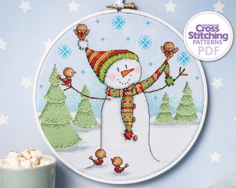 May include: A cross stitch pattern featuring a snowman wearing a green and red hat and scarf with birds perched on its arms. The pattern is in a white embroidery hoop against a light blue background with white stars.