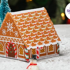 May include: A cross stitch pattern of a gingerbread house with white icing, candy canes, and a green wreath. The house is on a white background with a small snowman in the foreground. The text "The World of Cross Stitching Patterns PDF" is in the upper right corner of the image.