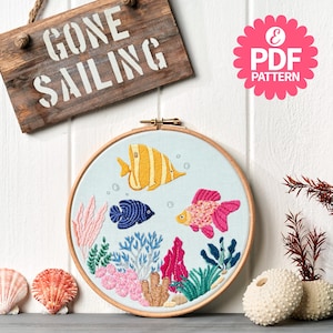 May include: Embroidered hoop art featuring an underwater scene with three colorful fish, coral, and plants. The artwork is set against a light blue background within a wooden hoop. A wooden sign above reads "GONE SAILING".