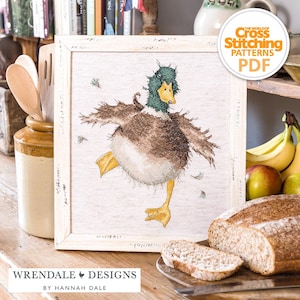 Peut inclure: Une broderie au point de croix encadrée représentant un canard dansant avec des détails bruns, verts et jaunes. L'œuvre est encadrée dans un cadre en bois blanc vieilli. Le texte "Wrendale Designs by Hannah Dale" est inclus.