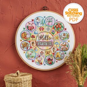 K&ouml;nnte beinhalten: Ein Kreuzstich-Stickrahmen mit einem farbenfrohen saisonalen Design. Das Design enth&auml;lt den Text "Hello September" und verschiedene kreisf&ouml;rmige Illustrationen von saisonalen Elementen wie K&uuml;rbissen, Blumen und einem Schneemann. Der Rahmen ist aus Holz und h&auml;ngt an einer roten Wand.
