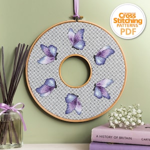 Puede incluir: Un bastidor de bordado de punto de cruz circular con un diseño de mariposas moradas y azules sobre un fondo geométrico en blanco y negro. El bastidor es de madera y tiene una cinta morada para colgar. El texto "The World of Cross Stitching Patterns PDF" es visible.