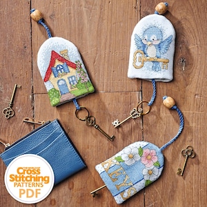 May include: A collection of cross-stitch key fobs and a blue leather cardholder are displayed on a wooden surface. The key fobs feature embroidered designs of a house, a bird with a key, and floral patterns. Antique-style keys are scattered around the items.