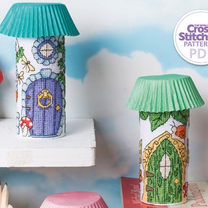 Op de afbeelding: Twee miniatuurhuisjes gemaakt van kartonnen buizen, versierd met kruissteekpatronen en getooid met groene cupcake-vormpjes. De huisjes staan op planken tegen een blauwe en witte wolkachtige achtergrond. De tekst "The World of Cross Stitching Patterns PDF" staat op een witte cirkel met een paarse rand.