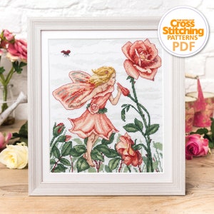Könnte beinhalten: Ein Kreuzstichmuster mit einer Fee mit rosa Flügeln und einem rosa Kleid, die eine rosa Rose riecht. Das Muster ist in einem weißen Rahmen gerahmt.